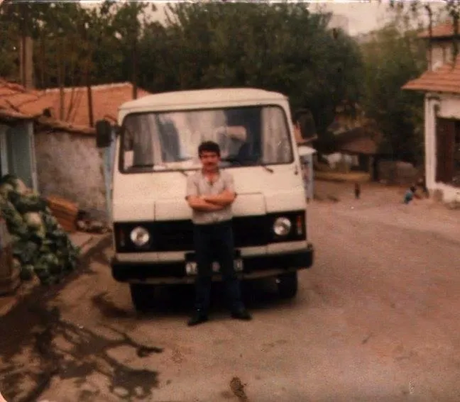 Eski bir fotoğrafta servis minibüsünün önünde kollarını kavuşturmuş duran şoför